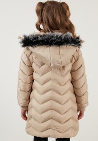 Beige polstret jakke med chevronmønster, faux fur kant ved hætten og ribbede manchetter. Glat tekstur og knælængde design.