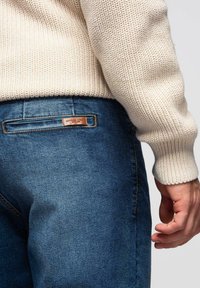 Pull en maille crème associé à un jean en denim bleu foncé, avec une étiquette en cuir sur la poche arrière, offrant un look décontracté et texturé.