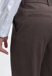 Λευκό μανίκι πουκάμισου με μανσέτα και χέρι δίπλα σε υφασμάτινο σκούρο καφέ παντελόνι με πίσω τσέπη με φάσα και πλαϊνή ραφή.