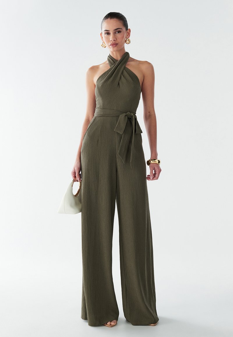 Olive green jumpsuit with a halter neck, tie waist, and wide-leg design. Textured fabric with a smooth finish. Accessories include a white bag.