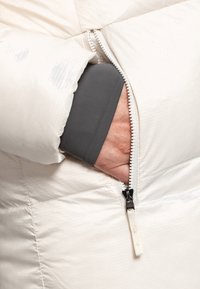 Pufferjack in licht crème met een ritssluiting pocket. Beschikt over textiel met structuur, zachte voering en een grijze manchet zichtbaar in de pocket.