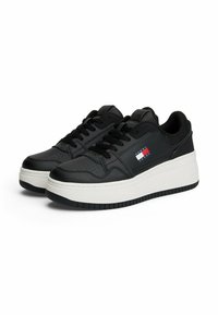 Sneaker sintetiche nere con suola bianca spessa, dotate di dettagli perforati, lacci neri e una piccola etichetta con logo nei colori rosso, bianco e blu.