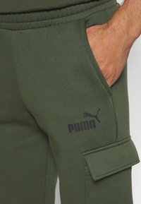 Pantalones de chándal verde oliva hechos de una tela suave, con un bolsillo lateral, cinturilla elástica y estampado en negro del logo de Puma en el muslo.