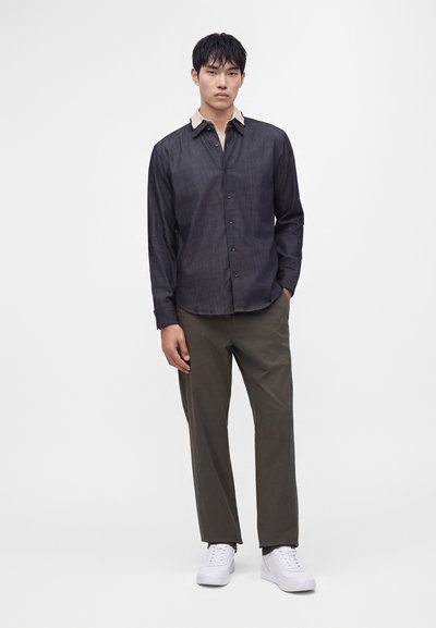 Homme debout portant une chemise sombre boutonnée avec un col beige, un pantalon vert olive et des baskets blanches sur fond blanc uni.