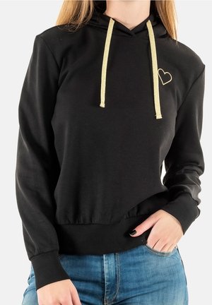 Sweat à capuche noir avec cordons dorés et petit cœur doré sur la poitrine, porté avec un jean bleu, main partiellement dans la poche.