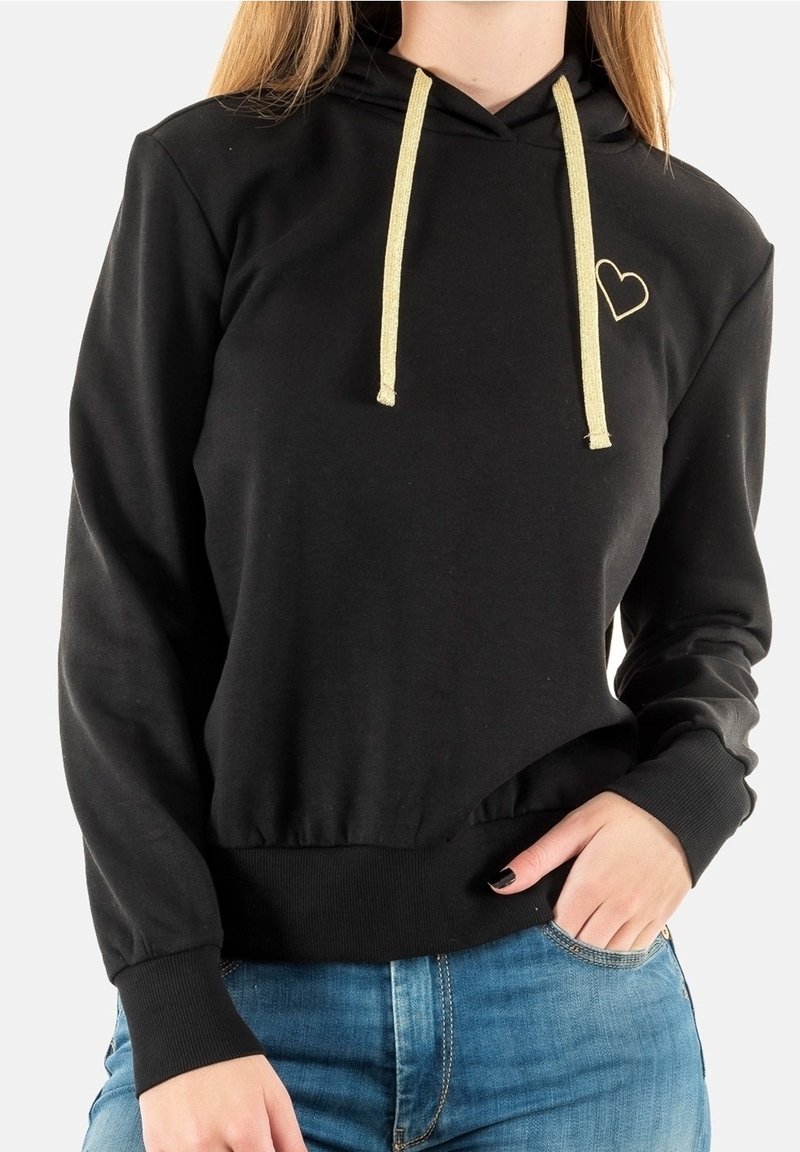 Sweat à capuche noir avec cordons dorés et petit cœur doré sur la poitrine, porté avec un jean bleu, main partiellement dans la poche.