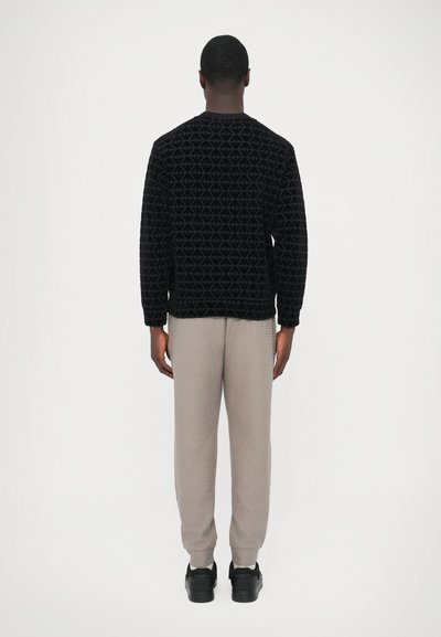 Čierny sveter s textúrou a geometrickým vzorom, v kombinácii s svetlosivými nohavicami a čiernou obuvou. Dizajn má dlhé rukávy a uvoľnený strih.