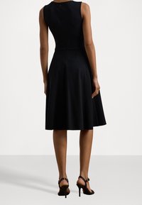 Svart ärmlös klänning med en åtsittande midja och utsvängd kjol, med en dragkedja i ryggen. Bärs tillsammans med svarta högklackade sandaler.