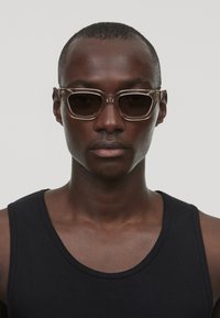 Lunettes de soleil avec une monture marron transparente et des verres foncés. Présentent un design géométrique avec des branches fines et des accents matériels subtils.
