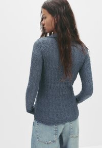 Langärmliges Oberteil aus strukturiertem dunkelblauem Stoff, mit taillierter Silhouette und Kragen, kombiniert mit hellblauen hoch geschnittenen Jeans.