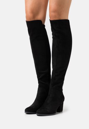 Botas de tacón alto de ante negro hasta la rodilla, usadas sobre piernas desnudas, de pie contra un fondo claro y liso.