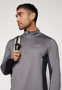 Pullover grigio Columbia con zip a mezza lunghezza in tessuto testurizzato, con accenti neri e logo sul petto. Il modello tiene una borsa a tracolla a strisce.