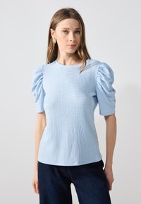 Ljusblå texturerad topp med korta puffärmar och rund halsringning. Bärs med mörkblå underdelar. Har en slät tygfinish.