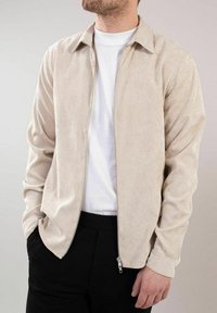 Beige kordfløyelsjakke med glidelås, krage og lange ermer, parret med en hvit T-skjorte og sorte bukser.