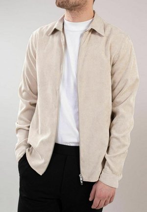 Beige kordfløyelsjakke med glidelås, krage og lange ermer, parret med en hvit T-skjorte og sorte bukser.