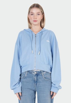 Junge Frau trägt eine hellblaue Kapuzenjacke mit Reißverschluss und Kordeln sowie blaue Jeans und steht vor einem einfarbig hellen Hintergrund.