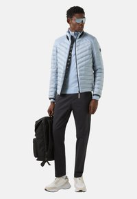 Lichtblauwe gewatteerde jas met een ritssluiting aan de voorkant, donkerblauwe broek, witte sneakers en een zwarte rugzak. Gestructureerde stof en sportief ontwerp.