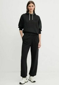 Donna in piedi che indossa un hoodie nero e pantaloni neri larghi con scarpe da ginnastica bianche, in posa contro uno sfondo chiaro e semplice.