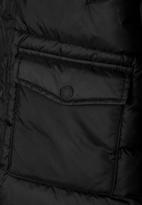 Juodas pūkinis švarkas su stačiakampiu kišeniu ir apvaliu juodu mygtuku. Medžiaga yra lygi, su siuvinėtu kvadratu raštu.