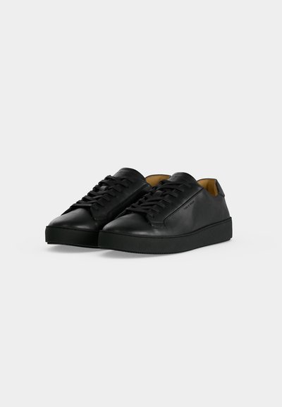 Ζεύγος μαύρα δερμάτινα sneakers χαμηλού ύψους με μαύρα κορδόνια και λαστιχένιες σόλες σε απλό ανοιχτό γκρι φόντο.