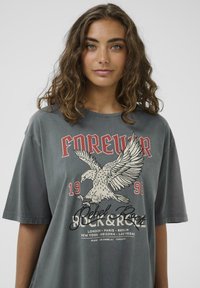 Grå oversized t-shirt med grafisk design af en ørn og rød tekst "FOREVER" samt "Rebel Rock" i sort, blød bomulds tekstur.
