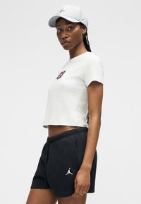 Γυναίκα που φοράει λευκό κοντό T-shirt με πολύχρωμο γραφικό, μαύρα σορτς Jordan και λευκό καπέλο Jordan, στέκεται μπροστά σε απλό φόντο.