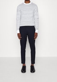 Bílý a šedý pruhovaný pletený svetr, slim-fit námořnické kalhoty a černé šněrovací boty, stojící na světlém pozadí.