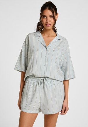 Vrouw draagt lichtblauw, loszittend, verticaal gestreept overhemd met knopen en bijpassende short met trekkoord, staand tegen een effen achtergrond.