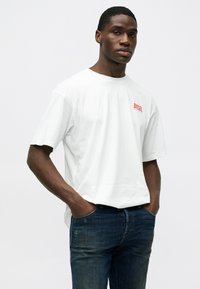 Camiseta de algodón blanca con un logo rojo de "DIESEL" en el frente, combinada con jeans de mezclilla azul oscuro. Diseño simple con mangas cortas.