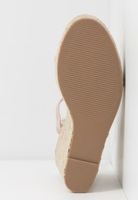 Semelle d’une sandale compensée beige avec une semelle en caoutchouc texturé et une plateforme en jute tissée, inclinée contre une surface d’angle blanche.