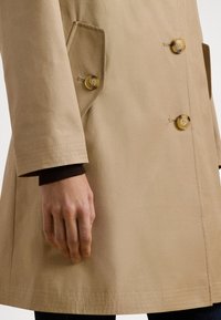 Beige trenchcoat med två knappar, en spetsig ficka och sydda muddar; tillverkad av slätt, lätt material.