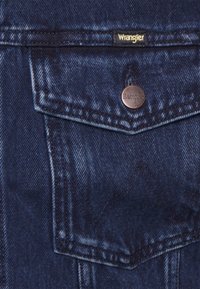 Tumma denimtakki, jossa edessä rintatasku pronssinvärisellä napilla ja Wrangler-logolla. Kankaan pinta on teksturoitu, ja siinä on hienovaraiset haalistumat ja ompeludetaljit.