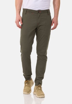 Muž nosící olivově zelené kalhoty slim fit, béžové tenisky a bílou košili zastrčenou dovnitř, stojí na bílém pozadí.