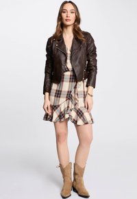 Veste de moto en cuir marron avec poches zippées, robe à carreaux crème avec ourlet à volants, et bottines en suède beige à bout pointu.