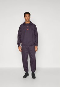 Marinblå sportsoverall i mjukt tyg, bestående av en huvtröja och avsmalnande byxor med röda logodetaljer. Svarta sneakers på fötterna.