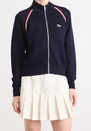 Pull zippé bleu marine avec bordure rouge et blanche aux épaules, associé à une jupe plissée blanche portée par une personne du cou à mi-cuisse.