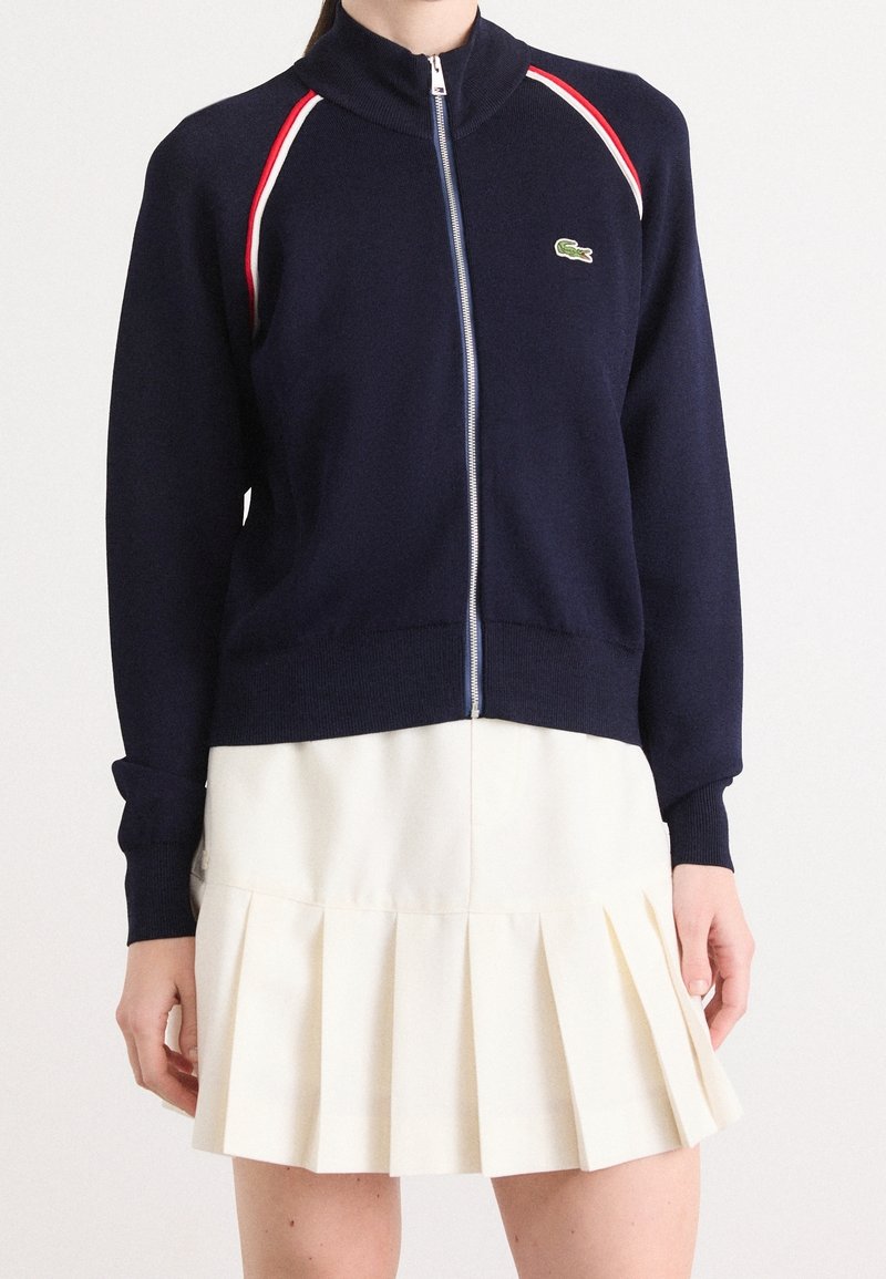 Pull zippé bleu marine avec bordure rouge et blanche aux épaules, associé à une jupe plissée blanche portée par une personne du cou à mi-cuisse.