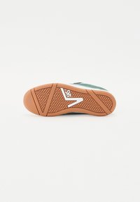 Semelle de chaussure en caoutchouc orange avec un motif hexagonal, arborant un logo blanc et une tige en daim vert avec des accents texturés.