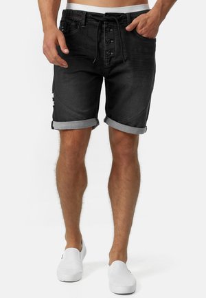 Shorts en denim noir avec un aspect légèrement déchiré, fermeture à cinq boutons, taille avec cordon de serrage et ourlet replié. Portés avec des chaussures blanches à enfiler.