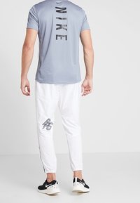 Muž v světlemodrém tričku Nike se svislým logem, bílých sportovních kalhotách s černým designem RS67 a černobílé tenisky Nike, stojí zády.