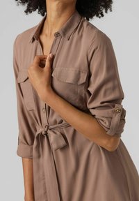 Donna che indossa un vestito mauve con bottoni, maniche arrotolate e vita legata, mentre tocca l'area del colletto con la mano destra.