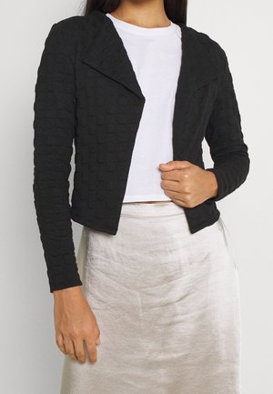Femme portant une veste courte noire texturée sur un haut blanc, associée à une jupe en satin de couleur claire, posant devant un fond uni.
