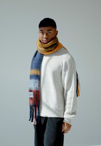 Écharpe en laine avec des rayures bleu, orange, gris et rouge, aux extrémités frangées, portée sur un pull à côtes blanc sur un fond neutre.