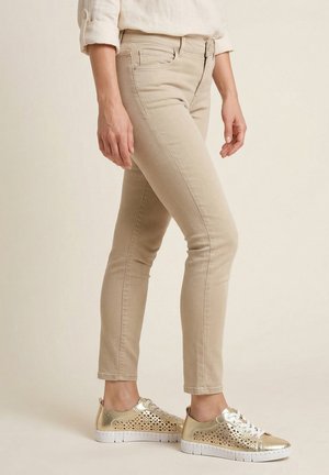 Vrouw draagt beige slim-fit broek en metallic gouden vetersneakers met witte zolen, staand tegen een effen achtergrond.