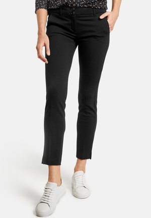 Femme portant un pantalon noir slim avec une ceinture à double bouton, associé à des baskets blanches et un haut noir à manches longues à motifs.