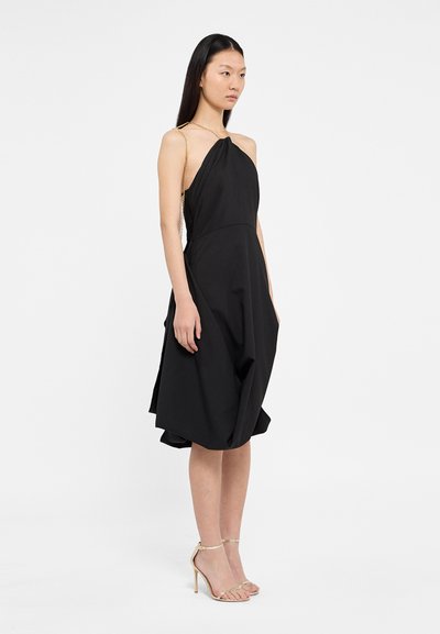 Črna brezrokavna obleka z drapirano zasnovo, ima zasukano korpus in zlate verižice, mehkega tekstila, midi dolžine, s prosto silhueto.