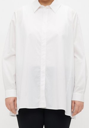 Persoon die een loszittend wit overhemd met lange mouwen en een klassieke kraag draagt, gecombineerd met een zwarte broek, staande tegen een effen achtergrond.