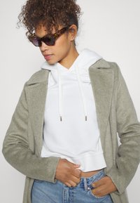 Cappotto grigio chiaro in misto lana su una felpa bianca con coulisse, abbinato a jeans blu. Gli accessori includono occhiali da sole con fantasia.
