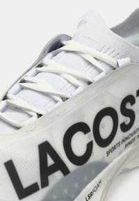 Κοντινός πλάνου ενός λευκού αθλητικού παπουτσιού με λευκές κορδόνες και μεγάλο μαύρο λογότυπο "LACOSTE" στο πλάι, που δείχνει μέρος της σόλας.