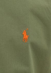 Orange broderad polo ryttare logotyp på olivgrön tyg med synlig textur och lätta veck.